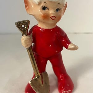 May include: A ceramic elf figurine wearing a red outfit and a matching red hat with a white brim. The elf has pointed ears, painted facial features, and is holding a small, metallic-colored shovel in its right hand.