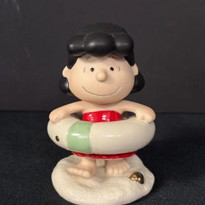 May include: A ceramic figurine of a cartoon character wearing a red swimsuit and a white and green life preserver. The figurine is standing on a sandy base with a gold accent.