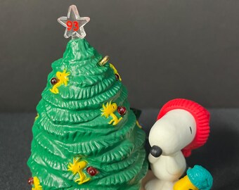 May include: A green Christmas tree with a star on top and red and yellow ornaments.  Snoopy is standing next to the tree wearing a red hat and scarf.  Snoopy's bird friend, Woodstock, is wearing a blue hat and is standing next to Snoopy.  The base of the tree is decorated with a red and white striped pattern.