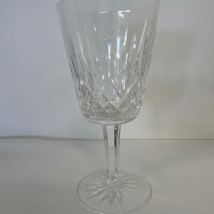 May include: A clear crystal wine glass with a long stem and a round base. The glass has a diamond-cut pattern on the bowl and a starburst design on the base. The glass is empty and sits on a white surface.