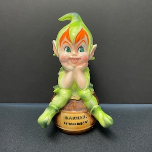 May include: A ceramic leprechaun figurine with green and orange details, sitting on a brown barrel. The barrel has the text "Barrel O' Money". The leprechaun has a smiling face and is resting its chin on its hands.