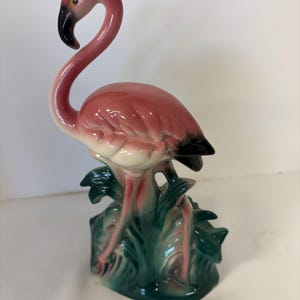 May include: A ceramic flamingo figurine featuring a glossy pink body with a curved neck, a black-tipped beak, and dark wing feathers. The bird stands atop a base sculpted to resemble green foliage and grass with a smooth, reflective finish.