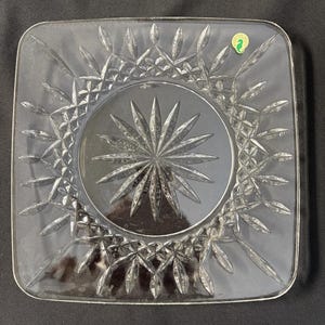 May include: A clear glass square plate with a starburst pattern in the center and a leafy design around the edge. The plate has a small green label with a seahorse logo.