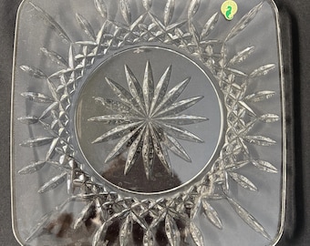 Könnte beinhalten: Ein quadratischer Teller aus klarem Glas mit einem Sternmuster in der Mitte und einem Blattdesign am Rand. Der Teller hat ein kleines grünes Etikett mit einem Seepferdchen-Logo.