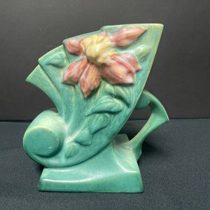 May include: A vintage art pottery vase in a unique shape, featuring a stylized daffodil design. The vase is a soft green color with a raised floral motif in shades of pink and cream. The vase has a distinctive handle and a geometric form.