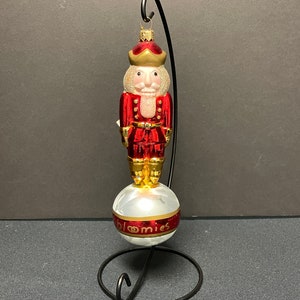 May include: A red and gold nutcracker Christmas ornament with a white face and a gold crown. The ornament is standing on a white and red ball with the word "bloomies" printed on it. The ornament is held by a black metal stand.