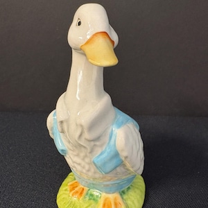 May include: A white ceramic duck figurine with a yellow beak and blue and white clothing. The duck is standing on a green base with orange feet.