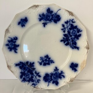 May include: A white ceramic plate with a scalloped edge featuring a deep blue floral pattern and gold-toned trim accents. The design includes clusters of flowers distributed across the surface, with a central floral motif and embossed details.