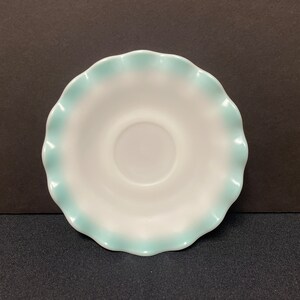 May include: A white saucer with a scalloped edge, trimmed in a light blue. The saucer has a circular indentation in the center. The design is simple and elegant, suitable for serving tea or coffee.