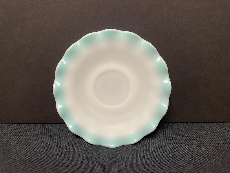 May include: A white saucer with a scalloped edge, trimmed in a light blue. The saucer has a circular indentation in the center. The design is simple and elegant, suitable for serving tea or coffee.