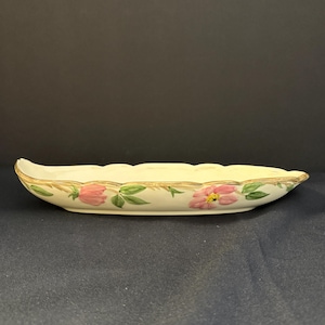 May include: A white ceramic serving dish with a scalloped edge and a floral design featuring pink roses and green leaves. The dish has a gold trim around the edge.