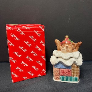 May include: A ceramic figurine of a brown bear wearing a white hat and a patchwork quilt. The bear is sitting on a base that is also decorated with a patchwork quilt. The figurine is in a red box with the letters "FF" repeated on the box.