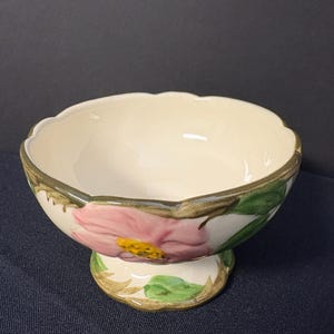 May include: A white ceramic bowl with a pink and green floral design. The bowl has a scalloped rim and a pedestal base.