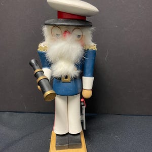 May include: A wooden nutcracker in the form of a sailor, wearing a blue jacket, white pants, and a white hat. The nutcracker has a white beard, glasses, and is holding a telescope. It stands on a wooden base and has gold trim.