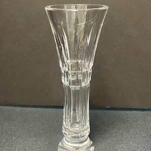 May include: A clear, cut-glass vase with a flared top and a detailed, geometric design. The vase has a round base and a slender, elongated body. The glass is transparent, allowing light to pass through, and is suitable for displaying flowers or as a decorative piece.