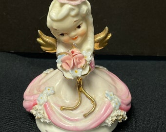 May include: A porcelain figurine of a girl angel with blonde hair, wearing a pink dress and holding a bouquet of pink roses. The figurine has gold wings and is sitting on a base with the words "TUESDAY'S CHILD" written on it.