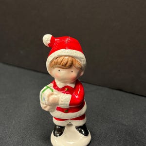 May include: A ceramic figurine of a child dressed in a Santa Claus outfit, holding a wrapped gift. The figurine has a red hat and suit with white trim, black boots, and a white base. A festive Christmas decoration.