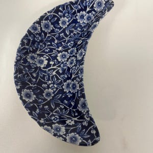 May include: A crescent-shaped ceramic dish featuring a dark blue and white floral pattern. The surface is covered in a dense, repeating design of small blossoms, leaves, and winding stems, creating a continuous botanical motif across the curved form.