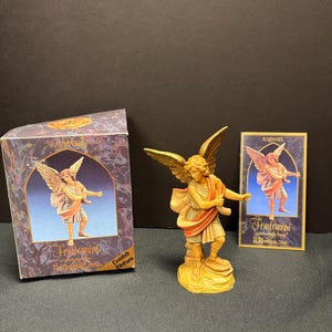 May include: A detailed figurine of an angel with outstretched wings, in shades of gold, tan, and red. The angel is standing on a base. A box and a card with the same angel image are also present.