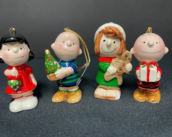 May include: Four ceramic Peanuts character Christmas ornaments. Lucy in a red dress, Charlie Brown with a Christmas tree, Sally with a gingerbread man, and Linus with a gift. Each figure has a gold string for hanging.