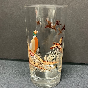 May include: A clear glass tumbler with a pheasant design. The glass features a detailed illustration of a pheasant and several flying birds in shades of brown, orange, and green. The design is set against a clear background.