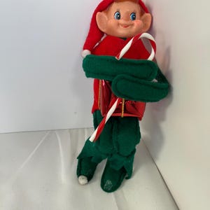 May include: A vintage Christmas elf doll with a red felt outfit, green felt boots and a green felt belt. The elf has a painted face with blue eyes and is holding a candy cane. A festive holiday decoration.
