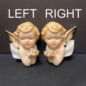 May include: Two porcelain cherub figurines, each holding a delicate pink flower. The cherubs have blonde curly hair, detailed wings, and are labeled "LEFT" and "RIGHT". The figurines are on a dark surface.