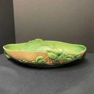 May include: A green and brown ceramic bowl with a leaf design. The bowl has a long, oval shape with a textured brown exterior and a smooth, green interior. Green leaves and vines are sculpted around the rim.