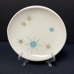 May include: A round, off-white ceramic plate with a speckled surface. The plate is decorated with a mid-century modern starburst design in yellow, blue, and gray. The plate is displayed on a clear plastic stand.