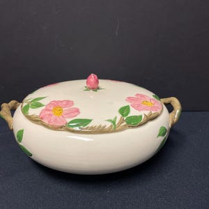 May include: A white ceramic casserole dish with a lid, featuring a floral design of pink roses with green leaves. The dish has two handles on the sides.