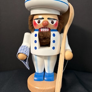 May include: A wooden chef nutcracker with a white hat, blue and white striped pants, and a blue and white checkered towel. The nutcracker is holding a wooden spoon.