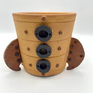 Könnte beinhalten: Ein handgefertigter Keramik-Pflanztopf mit einem einzigartigen Design. Der Topf ist hellbraun mit drei schwarzen kreisförmigen Akzenten und kleinen braunen Punkten. Er hat zwei braune, flügelartige Erweiterungen an den Seiten. Geeignet für kleine Pflanzen.