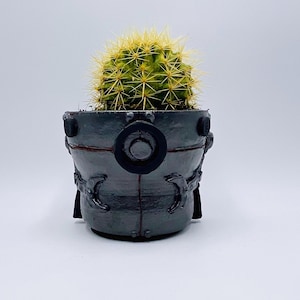 May include: A small, round cactus with yellow spines sits in a gray, industrial-style planter. The planter has a circular design element and curved accents. The cactus is a vibrant green color, contrasting with the gray pot.