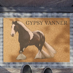 Può includere: Un cavallo Gypsy Vanner marrone e bianco con una lunga criniera e coda fluenti è raffigurato su uno zerbino color sabbia con il testo "GYPSY VANNER" stampato in nero. Lo zerbino ha un bordo a scacchi grigio e bianco.