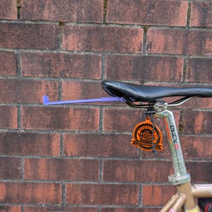Könnte beinhalten: Ein Fahrradsattel mit schwarzem Ledersitz und silberner Sattelstütze. Ein lila Stützarm erstreckt sich von der Sattelstütze. Ein orangefarbenes Etikett mit schwarzem Text hängt an der Sattelstütze. Der Hintergrund ist eine rote Backsteinmauer.