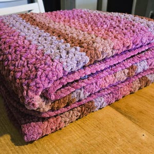 Puede incluir: Una manta plegada en tonos de rosa, morado y marrón. La manta está hecha de un material suave y texturizado y está apilada sobre una superficie de madera. Los colores hacen la transición en rayas a través de la manta.