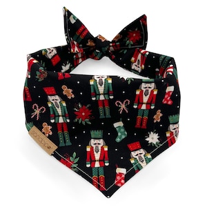 May include: Black dog bandana with a festive Christmas pattern. The bandana features nutcrackers, gingerbread men, poinsettias, and candy canes. The bandana is tied in a bow at the top.