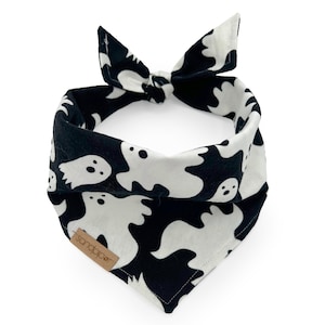 May include: Black and white Halloween-themed dog bandana with a repeating ghost pattern. The bandana is tied at the top with a bow. A small, brown tag with the word "sandpaper" is sewn on the bottom.