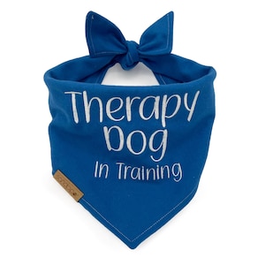 Puede incluir: Un pañuelo triangular azul real para mascotas con cierre de nudo y texto bordado en blanco que dice Therapy Dog In Training. Una pequeña etiqueta rectangular marrón con el nombre de la marca Sandpiper está unida al borde inferior izquierdo.