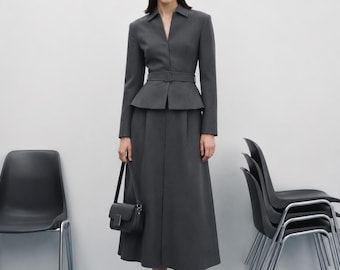 May include: A dark gray long-sleeved jacket with a notched collar, a belted waist, and a peplum hem, paired with a matching dark gray midi-length skirt. A small black rectangular shoulder bag and black pointed-toe heels complete the ensemble.