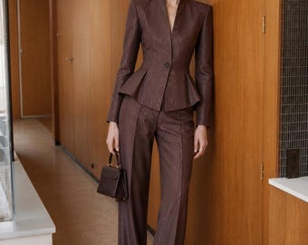 May include: A dark brown suit featuring a peplum jacket with a high collar and a single button closure, paired with matching wide-leg trousers. The fabric has a subtle wood grain texture. A small, structured dark brown handbag is held in one hand.