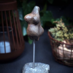 May include: A small, brown clay sculpture of a female figure on a gray stone base. The sculpture is mounted on a metal rod.
