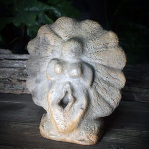 May include: A beige-colored, abstract sculpture with a textured surface. The sculpture features a stylized human form with a prominent open mouth and a radiating, shell-like design around the head. The piece is made of a stone-like material.