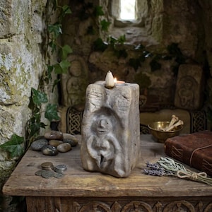 May include: A stone candle holder featuring a carved figure in a central niche, topped with a small metal plate holding a lit incense cone and a tea light candle. The textured, grey, block-like sculpture sits on a wooden surface with coins.