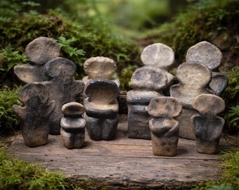May include: A collection of small, abstract sculptures in varying shades of gray and brown. The figures have rounded heads and stylized bodies, arranged on a weathered wooden surface. The background features lush green moss and foliage.
