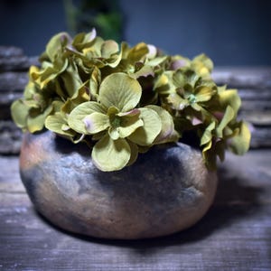 May include: A small, round, stone-like vase filled with artificial green and purple flowers. The vase is a mix of brown, gray, and tan colors. The flowers have a soft, textured appearance, and the arrangement sits on a wooden surface.