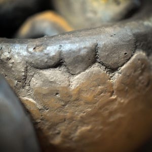 May include: A close-up view of a dark, textured ceramic vessel with a thick, uneven rim. The surface features a mottled appearance with shades of charcoal gray, deep brown, and muted bronze, showing a hand-formed, organic, and rugged construction.