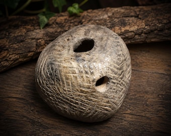 May include: A round, textured ceramic object with a mottled gray and beige finish. The surface features a dense, crisscross etched pattern throughout. It has two distinct circular openings, one positioned on the top and one on the side.