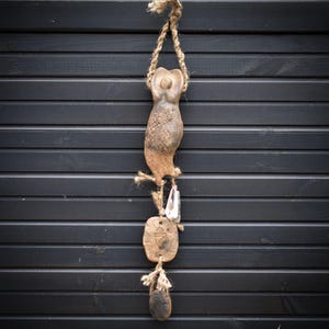 May include: A hanging, rustic-style wall decoration. The piece features a central, elongated, brown ceramic figure suspended from a natural rope. Below, two additional ceramic pieces hang, also connected by rope. The background is a dark, horizontal wooden surface.