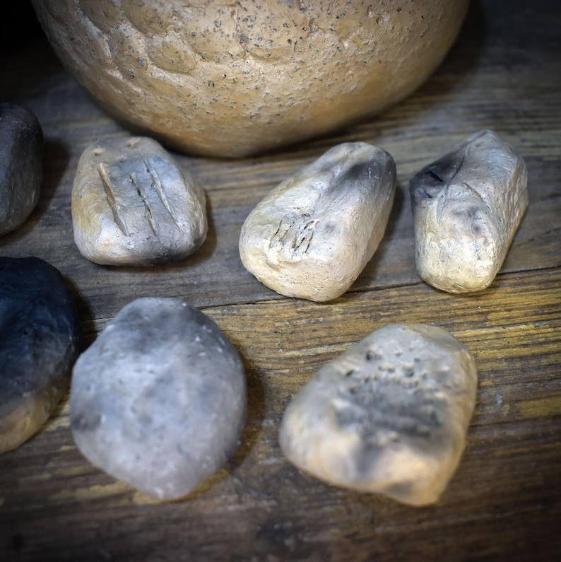 May include: A collection of small, irregularly shaped stones with a weathered, off-white and grey finish. Several of the stones feature carved, etched markings on their surfaces, while others remain smooth. The stones are arranged in a cluster.